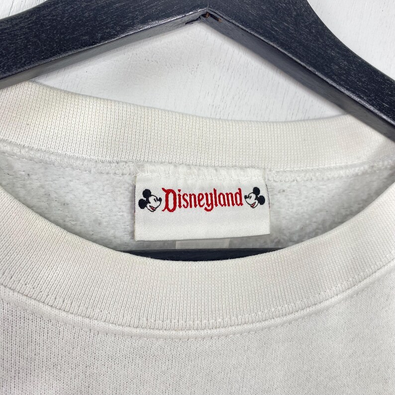 Puede incluir: Primer plano de un cuello de sudadera blanco con una etiqueta de Disneyland. La etiqueta presenta el logotipo de Disneyland en rojo y negro, con siluetas de Mickey Mouse. La sudadera tiene un cuello acanalado.