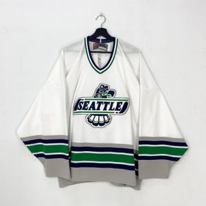 May include: White hockey jersey with the word "SEATTLE" in green and blue, featuring a stylized eagle logo. The jersey has green and blue stripes on the sleeves and collar. The jersey is a size 2XL.