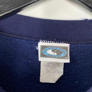 Puede incluir: Primer plano del cuello de una sudadera azul marino con una etiqueta. La etiqueta dice "TC Surf Designs Hawaii" con un s&iacute;mbolo de yin-yang. La etiqueta tambi&eacute;n incluye instrucciones de lavado. La sudadera est&aacute; hecha de un material suave.