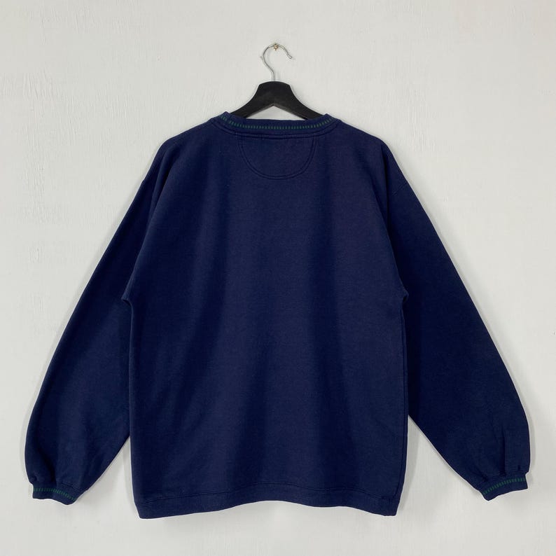 May include: Navy blue sweatshirt with a crew neck and long sleeves. The collar and cuffs have a green and navy striped trim. The back of the sweatshirt is visible, hanging from a black hanger against a white background.