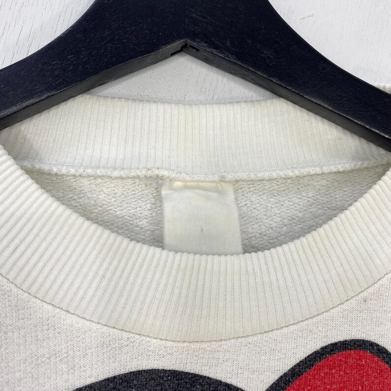 Puede incluir: Primer plano de una sudadera blanca de cuello redondo con cuello acanalado. La sudadera est&aacute; colgada de una percha negra. La parte inferior de la sudadera presenta un dise&ntilde;o rojo y negro.