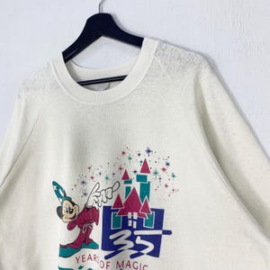 Pu&ograve; includere: Felpa color crema con una grafica di Topolino in abito da mago, un castello e la scritta "35 Years of Magic". Il design include accenti rossi, turchese e viola. La felpa &egrave; appesa a una gruccia nera.