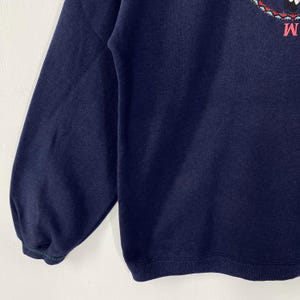 May include: Navy blue sweatshirt with an embroidered design near the neckline. The design includes a character and the letter "M". The sleeve cuffs have a green and navy blue trim.