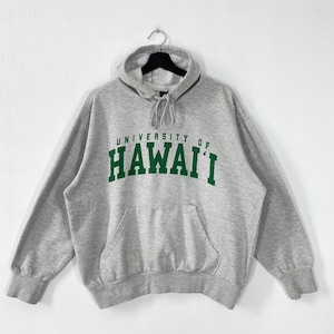 Könnte beinhalten: Grauer Kapuzenpullover mit dem grünen Schriftzug "UNIVERSITY OF HAWAII". Der Pullover hat eine Vordertasche und eine Kapuze mit Kordelzug. Das Kleidungsstück hängt an einem schwarzen Kleiderbügel vor weißem Hintergrund.