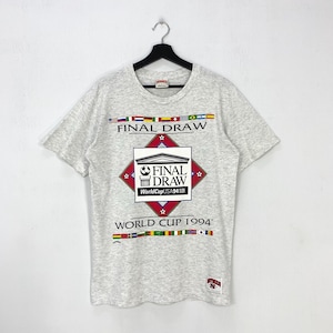 May include: Light gray t-shirt featuring the text "FINAL DRAW" above a central graphic with "FINAL DRAW WorldCup USA 94" and "WORLD CUP 1994" below. The shirt has a crew neck and short sleeves, with flags across the top and bottom.