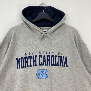 May include: A gray hoodie with a navy blue lined hood. The front of the hoodie features the words "UNIVERSITY OF NORTH CAROLINA" in navy blue lettering, with a light blue logo below. The hoodie is hanging on a black hanger.