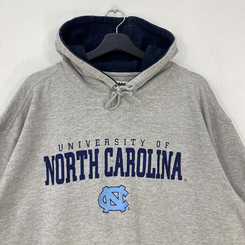 May include: A gray hoodie with a navy blue lined hood. The front of the hoodie features the words "UNIVERSITY OF NORTH CAROLINA" in navy blue lettering, with a light blue logo below. The hoodie is hanging on a black hanger.