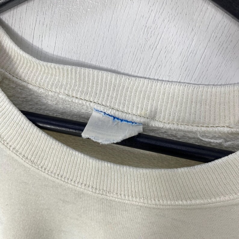 Pu&ograve; includere: Primo piano del colletto di una felpa color crema con dettagli a coste. Una piccola etichetta strappata con accenti blu &egrave; attaccata al colletto. Lo sfondo &egrave; una superficie bianca strutturata.