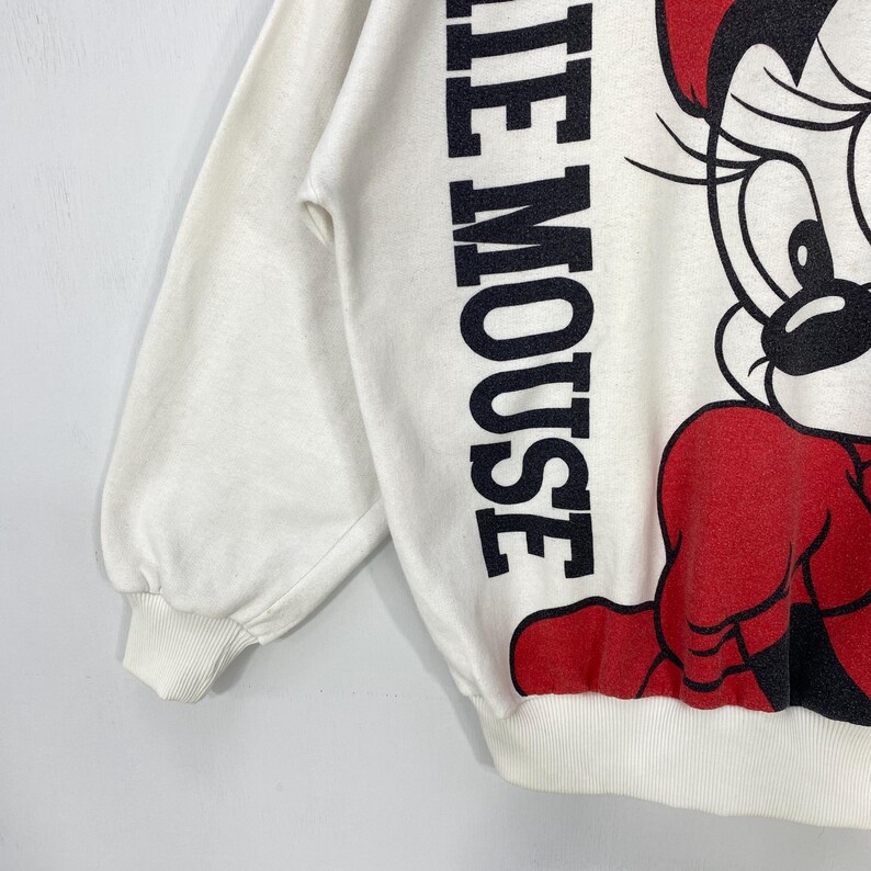 Puede incluir: Sudadera de color crema con un gran gr&aacute;fico de Minnie Mouse en rojo y negro. Las palabras "Minnie Mouse" est&aacute;n impresas verticalmente en negro en la manga izquierda. La sudadera tiene pu&ntilde;os y dobladillo acanalados.