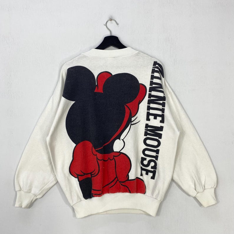 Puede incluir: Sudadera blanca con un gran gr&aacute;fico de Minnie Mouse en rojo y negro en la espalda. Las palabras "MINNIE MOUSE" est&aacute;n impresas verticalmente en la manga derecha en negro. La sudadera est&aacute; colgada de una percha negra.