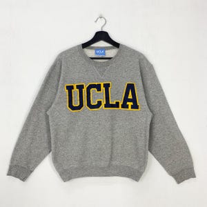 Puede incluir: Una sudadera gris de la UCLA con el logotipo de la UCLA en azul marino con un contorno dorado. La sudadera tiene cuello redondo y mangas largas. El logotipo de la UCLA está centrado en el pecho.