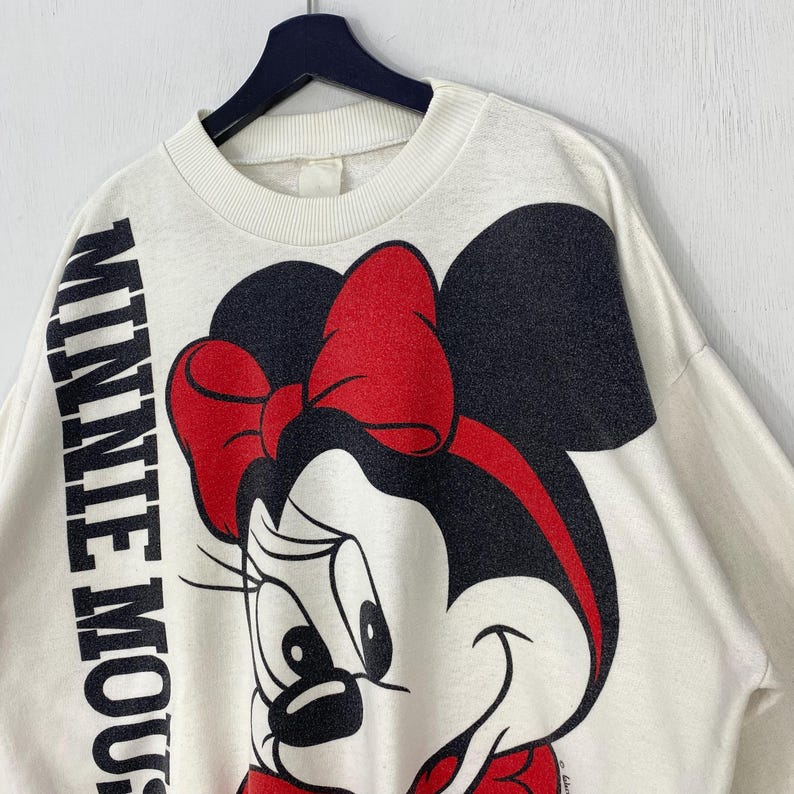 Puede incluir: Sudadera de color crema con un dise&ntilde;o de Minnie Mouse, con un lazo rojo y orejas negras. La palabra "MINNIE MOUSE" est&aacute; impresa verticalmente en negro en la manga izquierda. La prenda est&aacute; colgada en una percha negra.