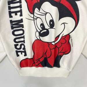 Puede incluir: Sudadera de color crema con un gran gr&aacute;fico de Minnie Mouse en rojo y negro. Las palabras "WHITE MOUSE" est&aacute;n impresas verticalmente en negro en el lado izquierdo. El logotipo de Disney es visible a la derecha.