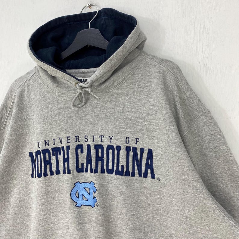 May include: A gray hooded sweatshirt with the words "UNIVERSITY OF NORTH CAROLINA" in navy blue across the chest. The sweatshirt has a light blue logo of a stylized letter "N" and "C" intertwined. The hood is lined with navy blue fabric.