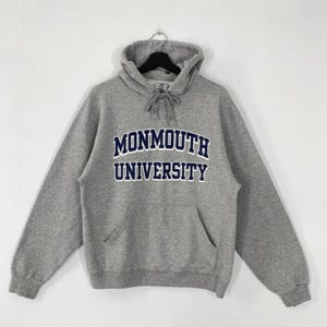 May include: A gray hooded sweatshirt with the text "MONMOUTH UNIVERSITY" in navy blue block letters. The sweatshirt has a front pocket and a drawstring at the hood. The garment is hanging on a black hanger.
