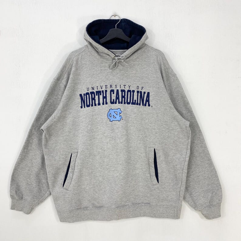 May include: A gray hooded sweatshirt with the text "UNIVERSITY OF NORTH CAROLINA" in navy blue. The sweatshirt has a navy blue lined hood and a front pocket. A light blue logo is below the text.