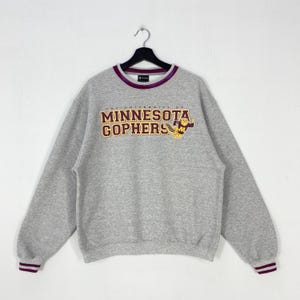 Puede incluir: Una sudadera gris jaspeado de manga larga con el texto "THE UNIVERSITY OF MINNESOTA GOPHERS" en letras granates y doradas, acompañada de un gráfico de mascota. El cuello redondo y los puños de las mangas tienen rayas granates y blancas.