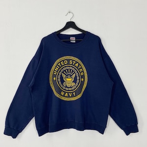 Könnte beinhalten: Marineblaues Sweatshirt mit einem großen, kreisförmigen Emblem mit der Aufschrift "UNITED STATES NAVY" in goldener Schrift. Das Emblem zeigt einen Adler und andere militärische Symbole. Das Sweatshirt hängt an einem schwarzen Kleiderbügel.