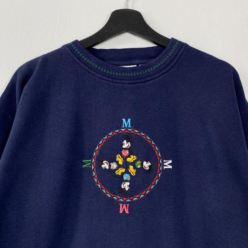May include: Navy blue sweatshirt with a crew neck and a green and white striped collar. The front features a colorful embroidered design of Mickey Mouse and friends within a circular pattern, with the letter "M" at each cardinal point.