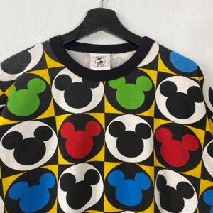 May include: A black crew neck sweatshirt featuring a repeating pattern of Mickey Mouse heads in black, red, green, and blue, set against a yellow and black checkered background. The collar is black, and the tag shows the Mickey Mouse logo.