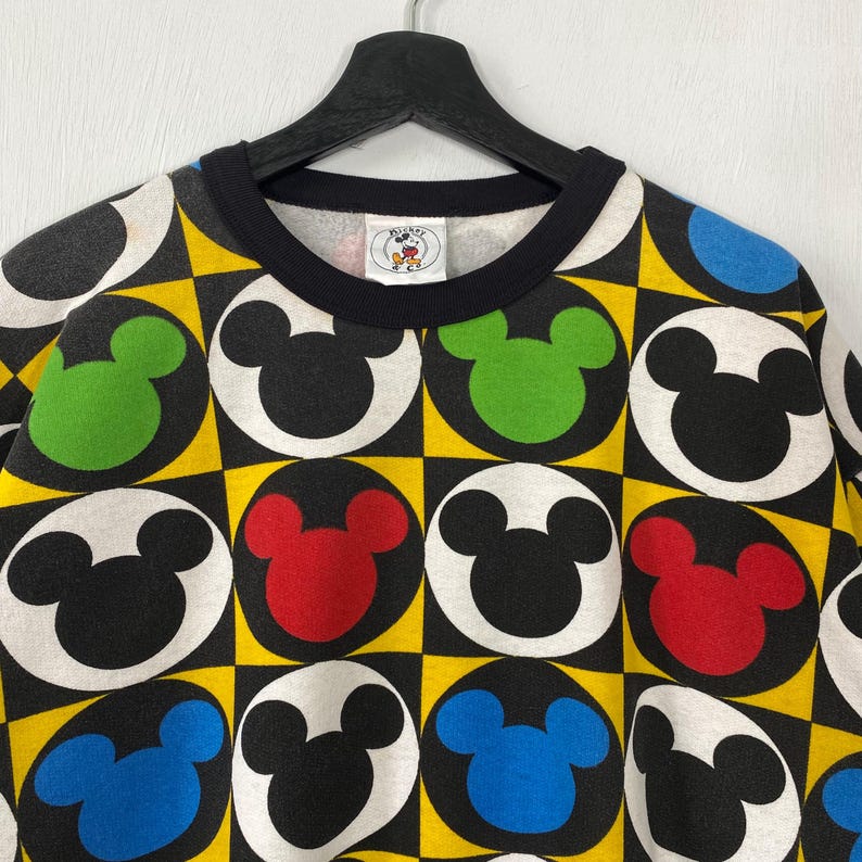 May include: A black crew neck sweatshirt featuring a repeating pattern of Mickey Mouse heads in black, red, green, and blue, set against a yellow and black checkered background. The collar is black, and the tag shows the Mickey Mouse logo.