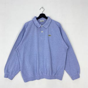 May include: A light blue long-sleeve polo shirt with a collar, a two-button placket, and a small green crocodile logo embroidered on the left chest. The shirt features a textured fabric and is displayed on a black plastic hanger.