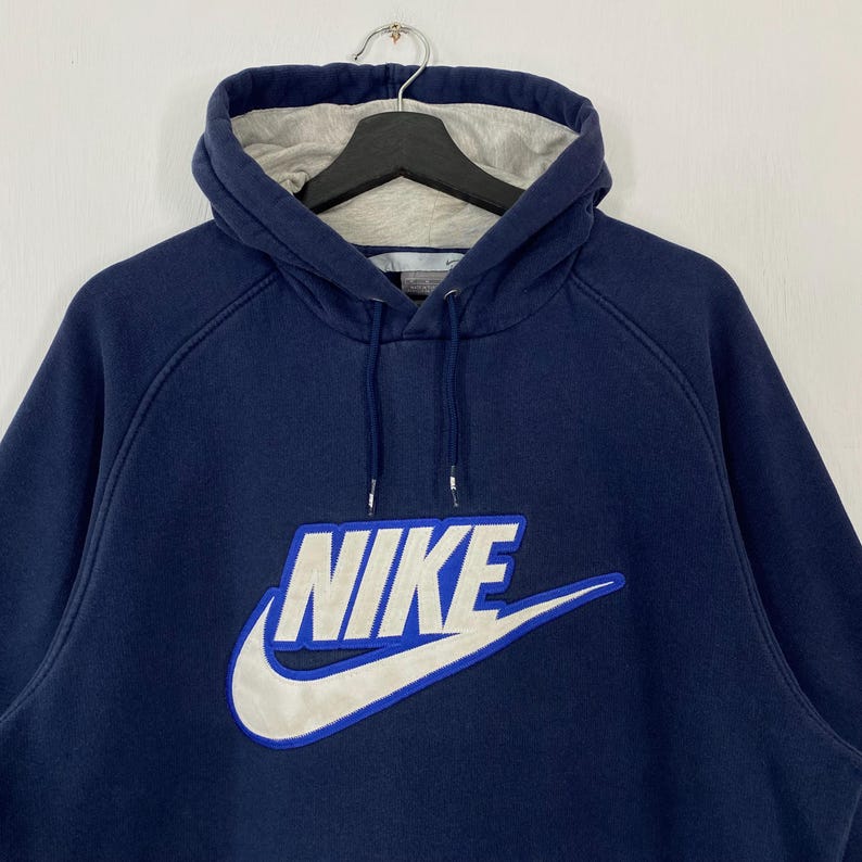 May include: Navy blue hoodie with a white and blue Nike logo on the front. The hoodie has a drawstring hood with a gray lining. The sweatshirt is hanging on a black hanger.
