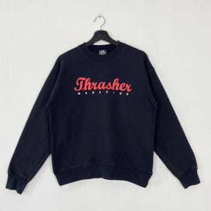 Puede incluir: Sudadera negra de cuello redondo con la palabra "Thrasher" en rojo en el pecho. Debajo de la palabra "Thrasher" está la palabra "Magazine" en letras blancas. La sudadera está colgada de una percha negra.