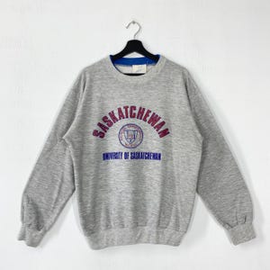 May include: A heather grey crewneck sweatshirt featuring the text SASKATCHEWAN in red arched lettering above a circular University of Saskatchewan crest and the text UNIVERSITY OF SASKATCHEWAN in blue. The collar has a blue inner trim.