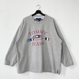 May include: A heather gray long-sleeve sweatshirt featuring the red embroidered text Tommy Hilfiger Jeans. The center graphic displays a row of international flags including the United Kingdom, United States, Canada, and Italy, all on a black hanger.