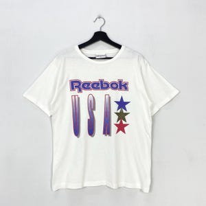 Op de afbeelding: Wit Reebok T-shirt met de merknaam in paars en blauw, en het woord "USA" in een gradiënt ontwerp. Voorzien van drie sterren in blauw, groen en rood. Het shirt hangt aan een zwarte hanger.