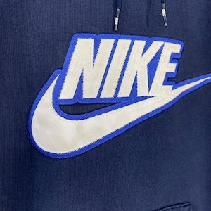 May include: Navy blue hoodie featuring a large white Nike logo with a blue outline. The Nike swoosh is also visible. The hoodie has a drawstring and a front pocket. The fabric appears to be a soft, comfortable material.