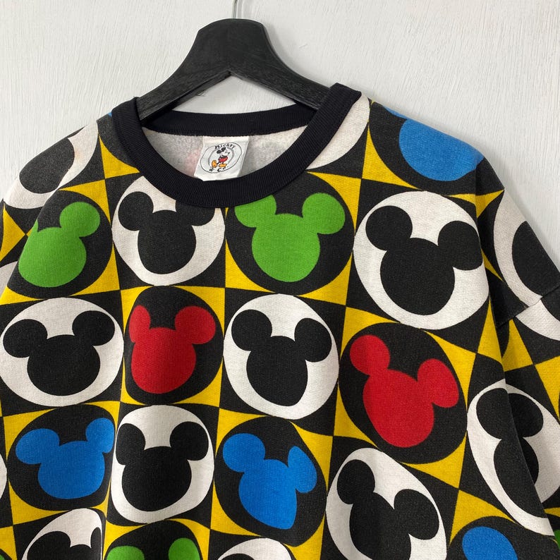 May include: A black crew neck sweatshirt with a colorful Mickey Mouse pattern. The design features Mickey Mouse heads in black, red, green, and blue circles set against a yellow and black checkered background. The collar is black.