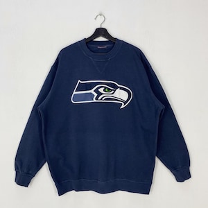 Puede incluir: Una sudadera azul marino de manga larga con cuello redondo y un gran logotipo de los Seattle Seahawks en el pecho. El logotipo muestra una cabeza de ave estilizada en blanco, azul marino y verde lima. Cuello, puños y dobladillo acanalados.