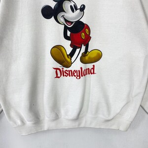 Puede incluir: Sudadera blanca con un gr&aacute;fico de Mickey Mouse con pantalones cortos rojos y zapatos amarillos. La palabra "Disneyland" est&aacute; impresa en rojo debajo. Una sudadera de estilo cl&aacute;sico.