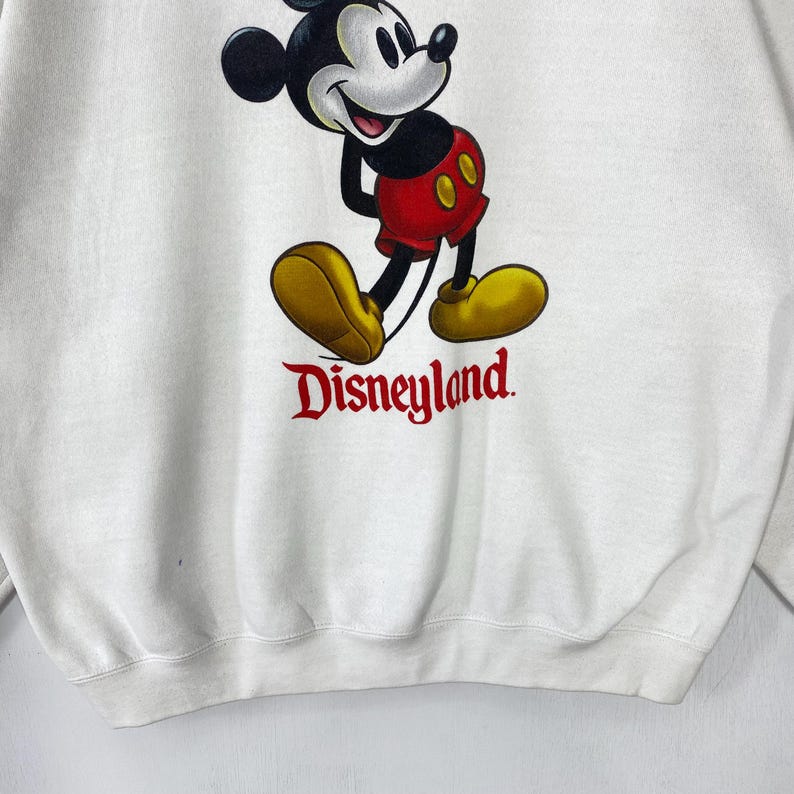 Puede incluir: Sudadera blanca con un gr&aacute;fico de Mickey Mouse con pantalones cortos rojos y zapatos amarillos. La palabra "Disneyland" est&aacute; impresa en rojo debajo. Una sudadera de estilo cl&aacute;sico.