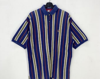 Könnte beinhalten: Blaues Poloshirt mit vertikalen Streifen in Rot, Weiß und Olivgrün. Das Shirt hat einen blauen Kragen und kurze Ärmel. Ein kleines Logo ist auf der linken Brust gestickt.