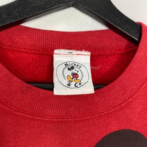 May include: A red crew neck sweatshirt with a Mickey & Co. label. The label shows a circular graphic of Mickey Mouse. The sweatshirt is made of a soft material and is likely vintage.