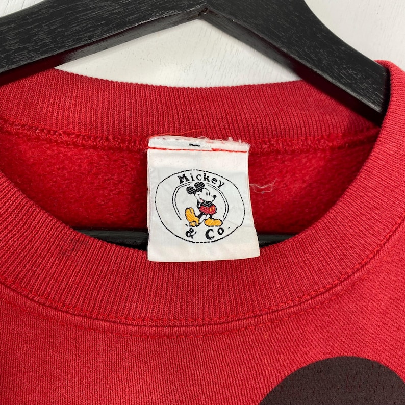 May include: A red crew neck sweatshirt with a Mickey & Co. label. The label shows a circular graphic of Mickey Mouse. The sweatshirt is made of a soft material and is likely vintage.