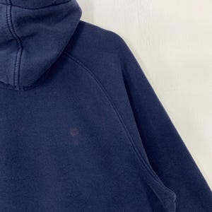 May include: Close-up of a navy blue hooded sweatshirt. The sweatshirt has a hood and a visible seam detail. A small stain is visible on the front of the garment. The background is a plain white surface.