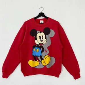 May include: Red crew neck sweatshirt with a large Mickey Mouse graphic. The cartoon character is in his classic outfit with yellow shoes, blue shorts, and black ears. A grey outline of Mickey Mouse is behind the character.