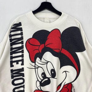 Puede incluir: Sudadera de color crema con un gran gr&aacute;fico de Minnie Mouse, con un lazo rojo y orejas negras. Las palabras "MINNIE MOUSE" est&aacute;n impresas verticalmente en negro en la manga izquierda. La sudadera est&aacute; colgada en una percha negra.