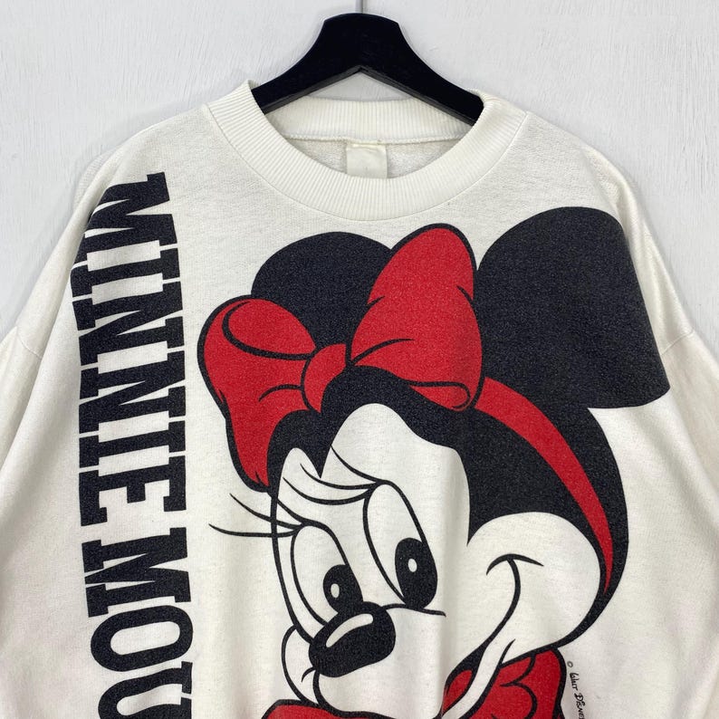 Puede incluir: Sudadera de color crema con un gran gr&aacute;fico de Minnie Mouse, con un lazo rojo y orejas negras. Las palabras "MINNIE MOUSE" est&aacute;n impresas verticalmente en negro en la manga izquierda. La sudadera est&aacute; colgada en una percha negra.