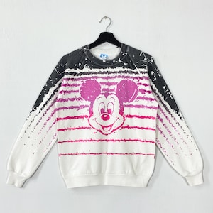 May include: White sweatshirt featuring a large Mickey Mouse graphic in pink. The design includes pink horizontal stripes and black accents on the shoulders. The sweatshirt is hanging on a black hanger.