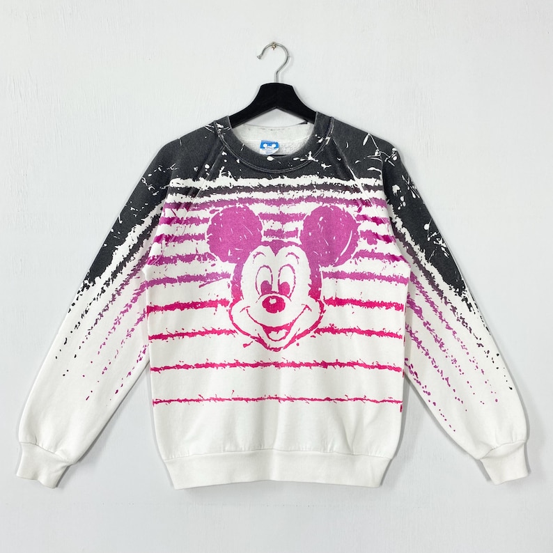 May include: White sweatshirt featuring a large Mickey Mouse graphic in pink. The design includes pink horizontal stripes and black accents on the shoulders. The sweatshirt is hanging on a black hanger.