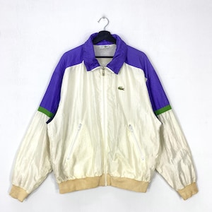 May include: A long-sleeve windbreaker jacket featuring a cream-coloured body with purple shoulders and sleeves, accented by thin green stripes. The jacket includes a full-length front zip, two zipped side pockets, and a small green crocodile logo.
