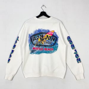 Op de afbeelding: Witte sweatshirt met een kleurrijk "Ron Jon Surf Shop" logo en een surfboard afbeelding. De mouwen tonen "Ron Jon" in blauw, groen en geel. De sweatshirt hangt aan een zwarte hanger.