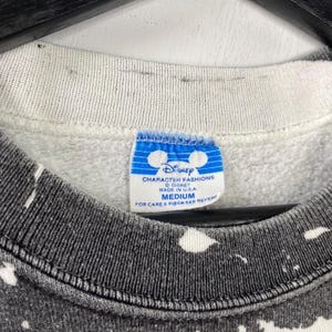 May include: Close-up of a vintage Disney Character Fashions sweatshirt. The tag reads "MEDIUM" and "MADE IN U.S.A." The sweatshirt has a gray and white speckled pattern. The collar is a light gray color.