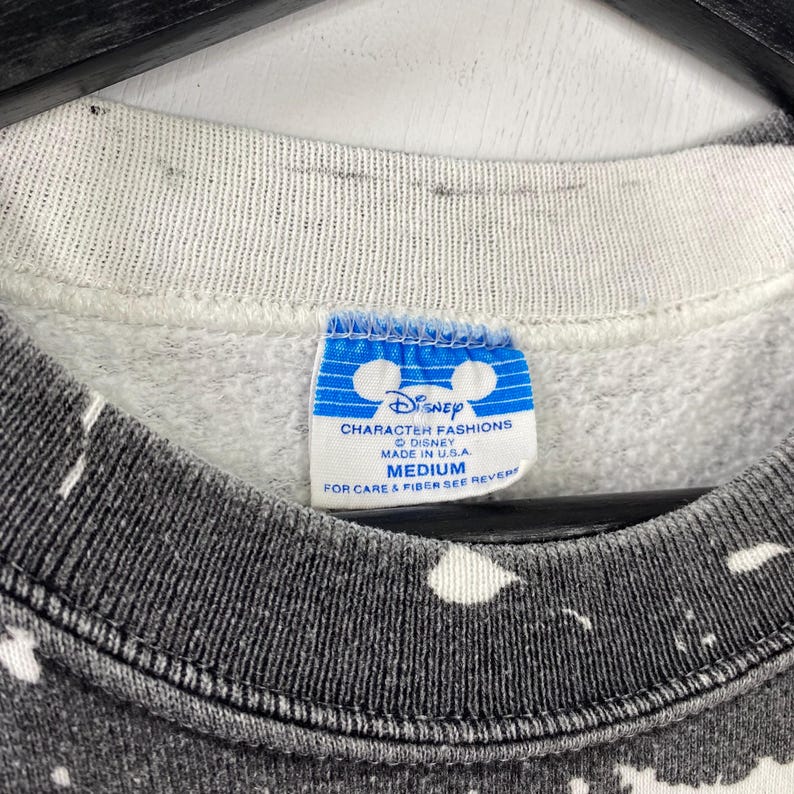 May include: Close-up of a vintage Disney Character Fashions sweatshirt. The tag reads "MEDIUM" and "MADE IN U.S.A." The sweatshirt has a gray and white speckled pattern. The collar is a light gray color.