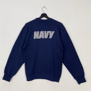 Könnte beinhalten: Ein marineblaues Sweatshirt mit dem Wort "NAVY" in weißen, diagonalen Streifen auf der Rückseite. Das Sweatshirt hat lange Ärmel und einen Rundhalsausschnitt und hängt an einem schwarzen Kleiderbügel.